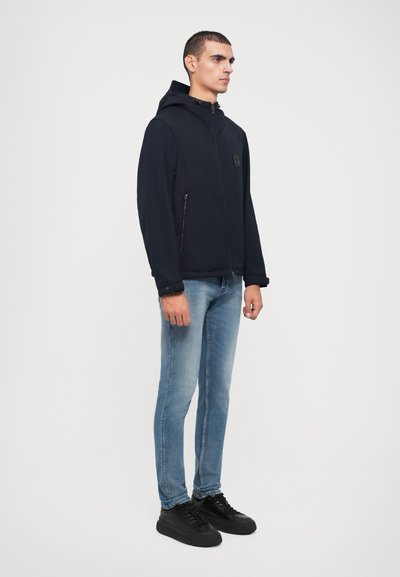Navy hooded jacket with zip closure, two side pockets, and a subtle logo patch. Worn over light blue denim jeans and black sneakers.