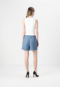 Top bianco senza maniche con un collo alto, abbinato a pantaloni corti blu con texture e orli sfrangiati, indossato con scarpe con tacco alte nere.