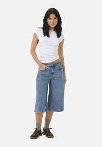 Top bianco a maniche corte, aderente. Culotte in denim blu morbido, lunghezza fino al ginocchio. Il modello indossa scarpe scure con suola spessa e calze grigie a coste.