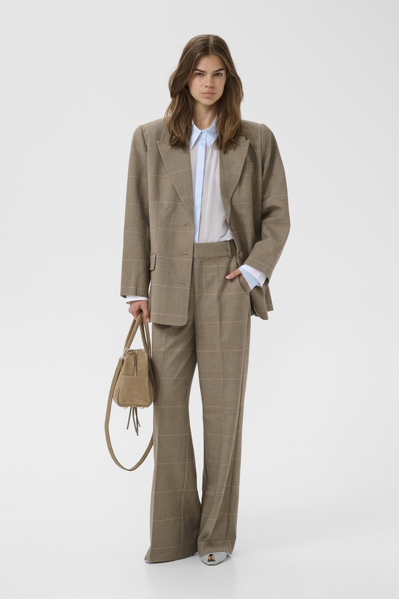 Jeune femme portant un costume oversize à carreaux beige avec un pantalon à jambes larges et un chemisier blanc, tenant un sac à main beige et des chaussures blanches.