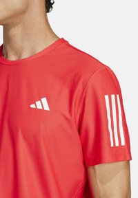 Camisa deportiva roja hecha de tejido texturizado, con un logo blanco en el lado izquierdo del pecho y tres rayas blancas verticales en la manga derecha.