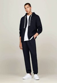 Mørkeblå fleece hoodie med hvide snore, parret med matchende joggerbukser og en hvid T-shirt. Modellen står i hvide sneakers.