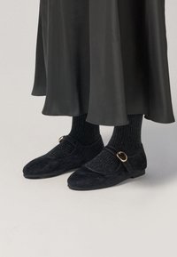 Zapatos Mary Jane de ante negro con un detalle de hebilla redonda, combinados con calcetas oscuras acanaladas, debajo de una falda negra fluida.