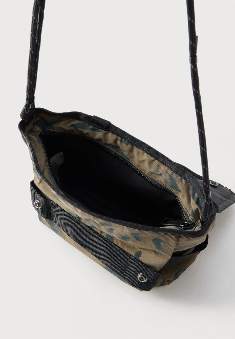 Pequeña bolsa bandolera con estampado de camuflaje, correa negra ajustable y parte superior abierta que muestra un compartimento interior con cremallera.