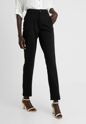 Persoon draagt zwarte, op maat gemaakte enkellange broek, wit overhemd met opgerolde mouwen en crème enkellaarzen met blokhak en enkelbandje.