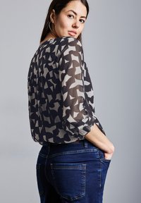 Průhledná černá blůza s světle šedým abstraktním vzorem, kulatým výstřihem a tříčtvrtečními rukávy, kombinovaná s tmavými džínami, viditelná přední kapsa.