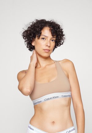 Bralette color tan con spalline sottili e una fascia bianca con la scritta "Calvin Klein" in testo nero. Texture liscia, design minimale, copertura media.