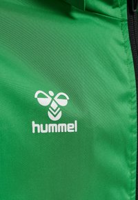 Veste de sport verte en nylon lisse avec une fermeture éclair noire. Équipée d'un logo "hummel" blanc et d'un graphique stylisé d'abeille.