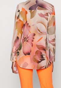 Blouse à manches longues, imprimé floral, en tissu transparent avec des nuances de rose, orange et vert. Comprend un ourlet arrondi et des poignets boutonnés. Associée à un pantalon orange vif.