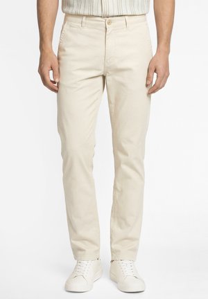 Mężczyzna w beżowych, dopasowanych spodniach, białych niskich trampkach i jasnej, w paski koszuli, stojący na jednolitym tle.