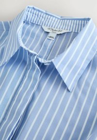 Chemise col bleu clair avec des rayures blanches verticales. Fabriquée en tissu doux, elle comprend une étiquette indiquant la marque et les instructions de lavage.