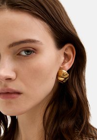 Goldfarbene Ohrstecker mit einem texturierten, wellenförmigen Design, das eine dreidimensionale Form und eine polierte Oberfläche aufweist, getragen am rechten Ohr.