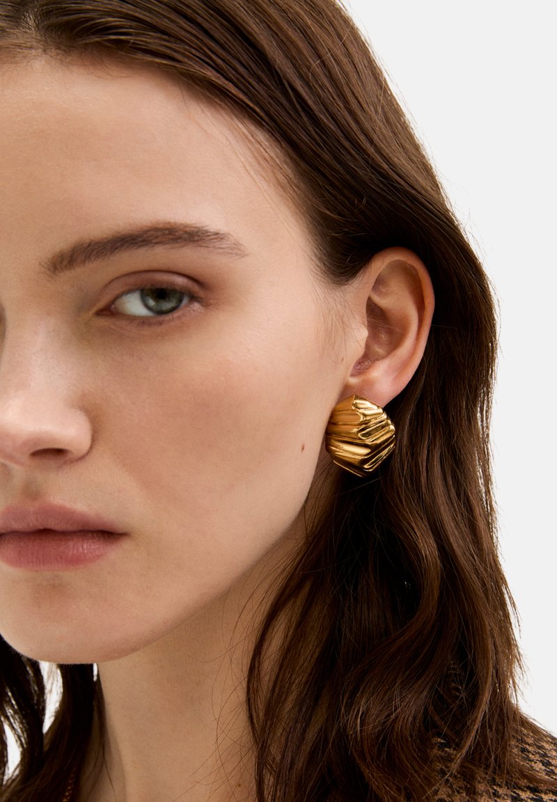 Goldfarbene Ohrstecker mit einem texturierten, wellenförmigen Design, das eine dreidimensionale Form und eine polierte Oberfläche aufweist, getragen am rechten Ohr.