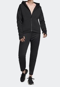 Svart zip-up huvtröja med långa ärmar och en ficka fram, tillsammans med matchande åtsittande joggers. Tyget verkar mjukt och bekvämt.