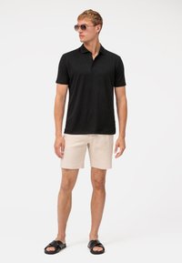 Schwarzes Polo-Shirt mit kurzen Ärmeln, beigefarbene Shorts und schwarze Sandalen. Das Modell trägt Sonnenbrillen und steht vor einem schlichten weißen Hintergrund.