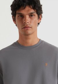 Graues Kurzarm-T-Shirt aus weichem Baumwollmaterial, mit Rundhalsausschnitt und einem kleinen orangefarbenen Logo auf der linken Brust. Glatte Textur.