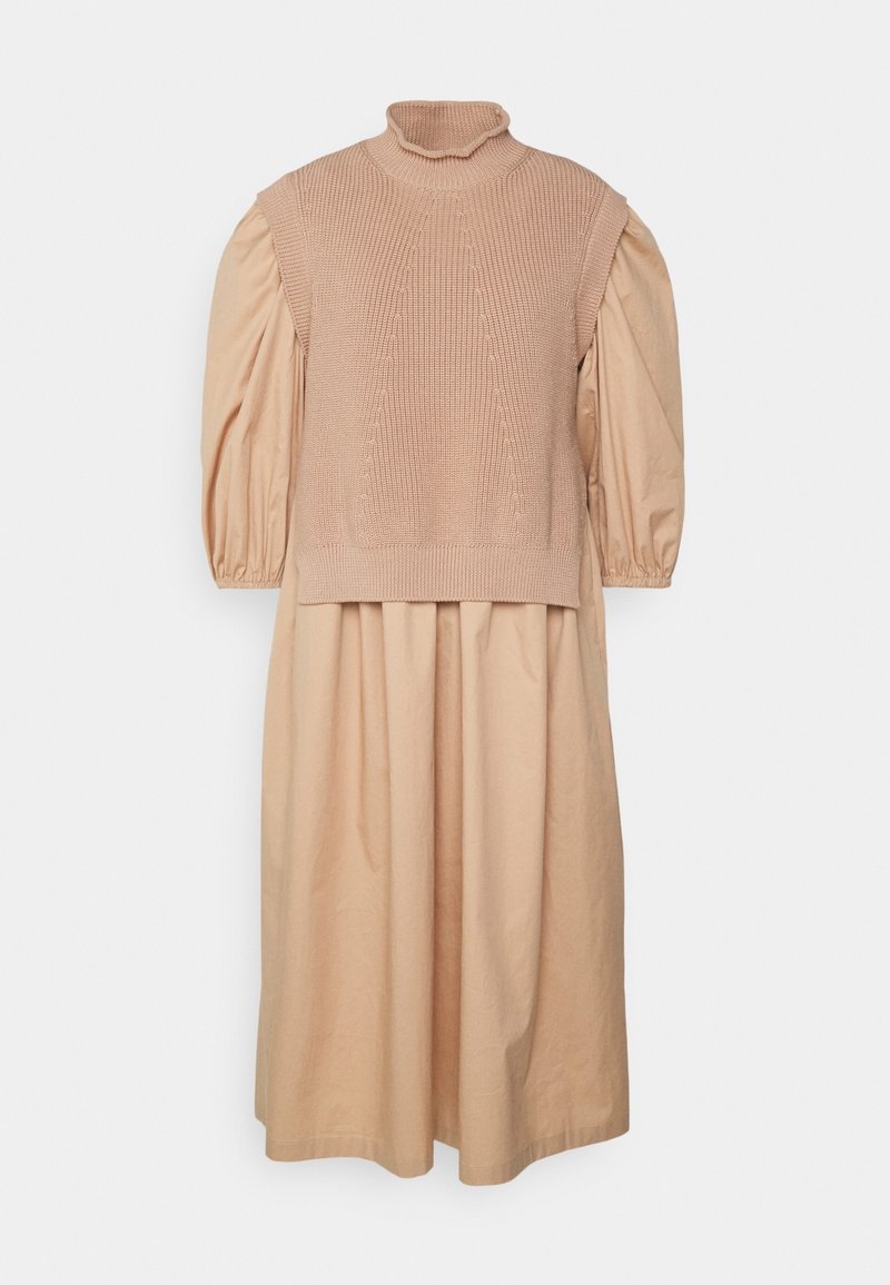 Robe longue beige à manches bouffantes associée à un pull en maille beige sans manches à col roulé superposé.