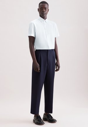 Mand iført en kortærmet hvid skjorte med knapper, marineblå bukser og sorte pæne sko, stående foran en ensfarvet baggrund.