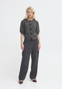 Cardigan grigio a maglia con una finitura testurizzata, caratterizzato da maniche a sbuffo e bottoni floreali neri. Abbinato a pantaloni grigi scuri sartoriali.