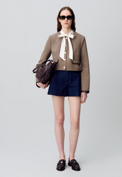 Veste cropped à carreaux bruns avec un nœud de cou blanc, shorts en denim et mocassins foncés. Le modèle porte un sac à main en cuir noir et des lunettes de soleil.