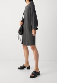 Abito in maglia grigio oversize con maniche lunghe, abbinato a sandali neri in pelle con tacco a blocco. La modella tiene una piccola borsa nera.