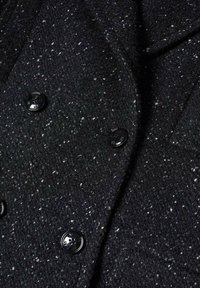 Manteau en laine noire texturée avec des mouchetures blanches et quatre grands boutons noirs disposés en style croisé.