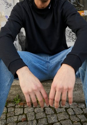 Osoba w czarnej bluzie i niebieskich dżinsach siedząca na kamiennych schodach z rękami luźno zwisającymi na kolanach, graffiti w tle.
