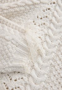 Tissu tricoté blanc avec des motifs complexes à torsades et en dentelle, avec un délicat ruban de dentelle noué au centre.