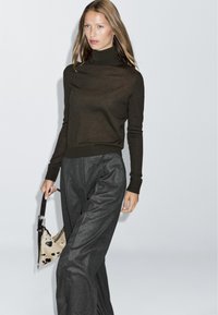 Brązowy sweter z golfem w połączeniu z luźnymi szarymi spodniami; trzyma beżową torebkę w czarne plamki. Gładki materiał, minimalistyczny design.