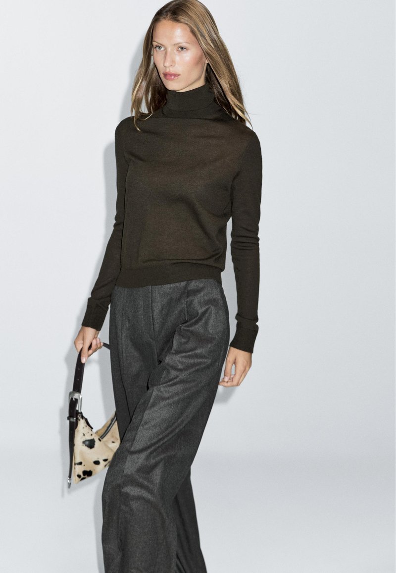 Brązowy sweter z golfem w połączeniu z luźnymi szarymi spodniami; trzyma beżową torebkę w czarne plamki. Gładki materiał, minimalistyczny design.