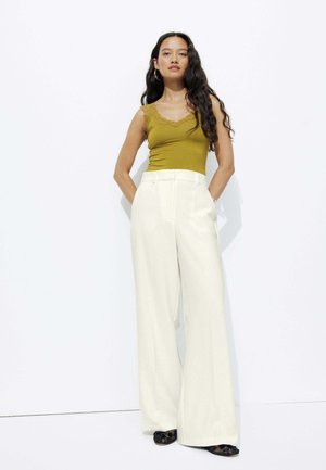 Débardeur vert olive avec dentelle, associé à un pantalon blanc taille haute et large. Le modèle se tient devant un fond uni.