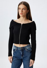 Czarny prążkowany sweter crop z dekoltem typu off-the-shoulder i zamkiem z przodu. Zestawiony z niebieskimi dżinsami o wysokim stanie.