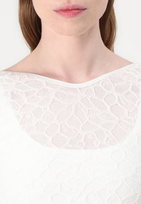 Top alb cu un model texturat și un design asemănător plaselor. Decolteu larg și țesătură netedă cu detalii subtile de suprapunere.