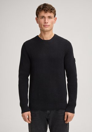 Junger Mann mit kurzen lockigen Haaren, der einen schwarzen strukturierten Strickpullover und dunkle Hosen trägt, vor einem schlichten hellen Hintergrund stehend.