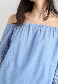 Light blue off-the-shoulder top made of soft fabric, featuring loose sleeves with eyelet embroidery at the hem. Gathered neckline detail.