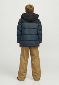 Pūka jaka ar melnu kapuci un torso, tumši zilo piedurkņu un horizontālu quiltu. Valkāta ar gaiši brūnām platām biksēm.