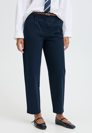 Personne portant un pantalon droit bleu marine, une chemise blanche avec une cravate rayée, une ceinture marron et des chaussures Mary Jane noires, debout devant un fond blanc.