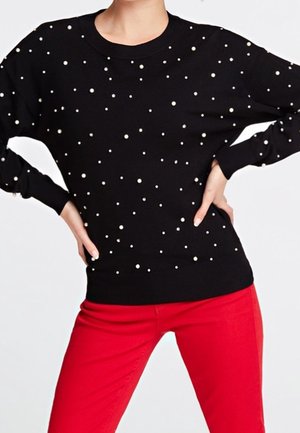 Personne portant un pull noir orné de perles blanches dispersées et un pantalon rouge vif, les mains posées sur les hanches devant un fond uni.