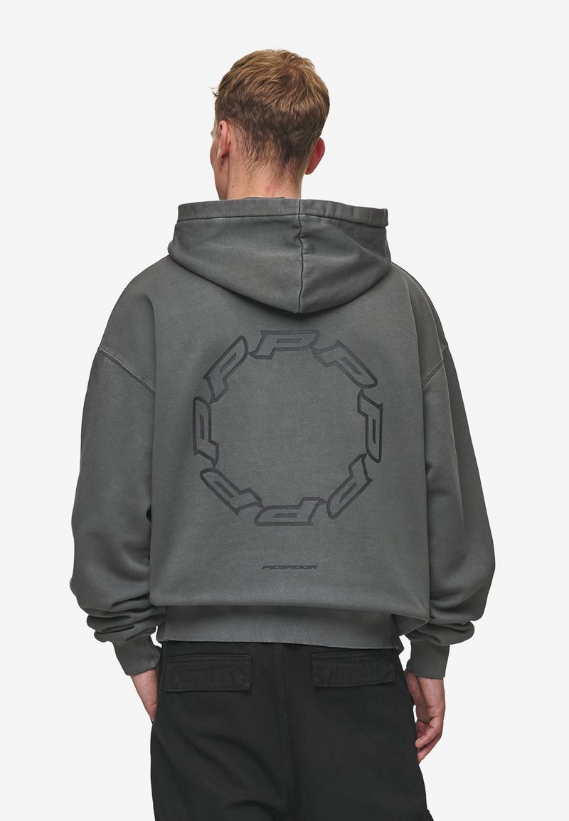 Homme portant un sweat à capuche gris foncé avec un logo circulaire "PRP" dans le dos, associé à un pantalon noir, tourné de dos sur un fond uni.
