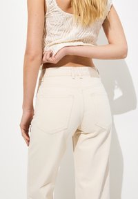 Jeans de talle alto beige con cintura ajustada, piernas rectas y dos bolsillos traseros. Tejido suave con detalles de costura visibles.