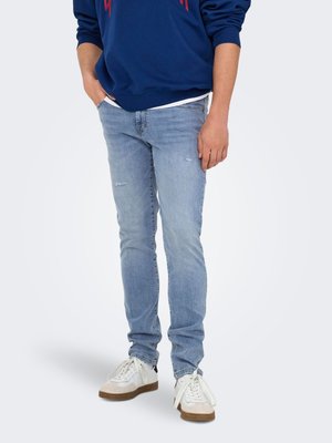 Homme portant un jean skinny bleu clair, des baskets blanches et un sweat bleu, les mains dans les poches, debout sur un fond blanc.