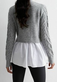 Grauer Strickcardigan mit Kabelstrickärmeln, cropped Design und geripptem Saum, kombiniert mit einer weißen Bluse aus fließendem, strukturiertem Stoff.