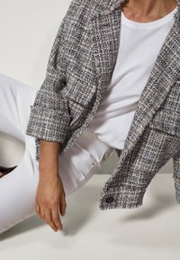 Tweedowy blazer o teksturze w kolorze niebieskim i kremowym, z postrzępionymi brzegami, noszony na białym T-shircie. W zestawieniu z białymi dopasowanymi spodniami.