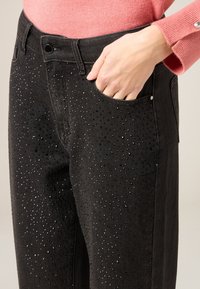 Mano derecha en el bolsillo del pantalón negro decorado con pequeñas tachuelas negras y plateadas, combinado con una camiseta rosa de manga larga con puño abotonado.