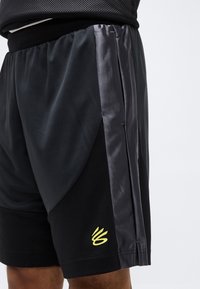 Κοντινό πλάνο μαύρου και σκούρου γκρι αθλητικού σορτς με ελαστική ζώνη  μέσης και κίτρινο στυλιζαρισμένο λογότυπο κοντά στο τελείωμα σε ένα άτομο.