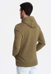 Sudadera verde oliva con capucha, diseño ajustado, puños y dobladillo acanalados, y textura suave. Vista desde la parte de atrás, sin marcas ni patrones visibles.