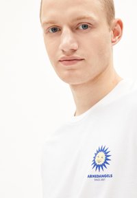Weißes Baumwoll-T-Shirt mit einem blauen Sonnenmotiv und dem Text "ARMEDANGELS seit 2007" auf der linken Brustseite. Glatte Textur, Rundhalsausschnitt.