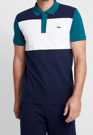 Mężczyzna w koszulce polo z krótkim rękawem w kolorze teal, w poziome niebieskie i białe paski, z małym zielonym logo krokodyla na piersiach.