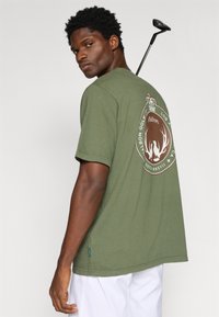 Olīvzaļš kokvilnas T-krekls ar lielu emblēmu aizmugurē, kurā attēlots kronis un ragi. Vienkāršs apaļais kakls un īsas piedurknes.