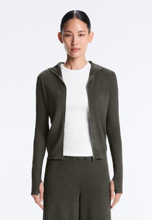 Woman wearing a dark green zip-up hoodie with ribbed sleeves over a white shirt, paired with matching dark green pants against a plain background.